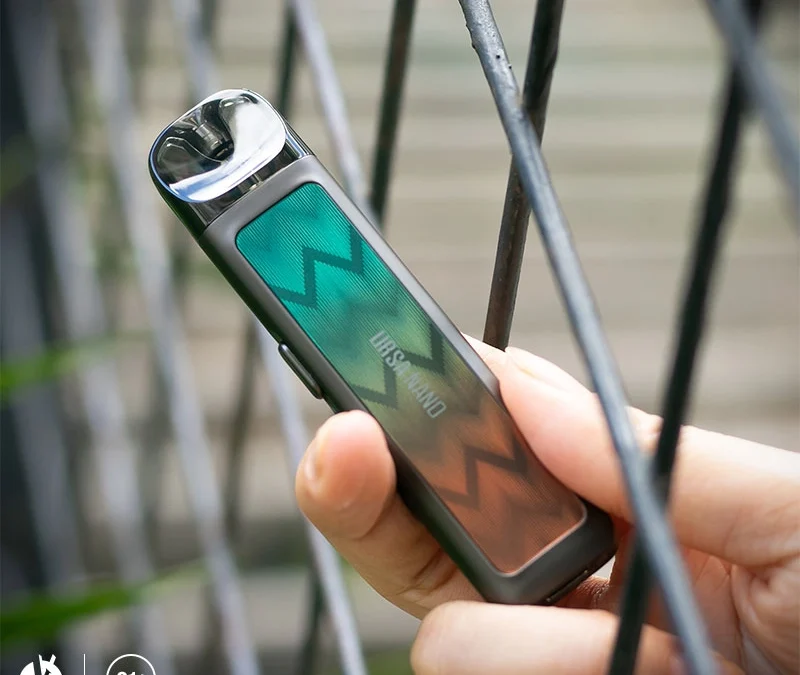 A hand holding an original Lost Vape URSA NANO beside a metal railing, with the model name visible.